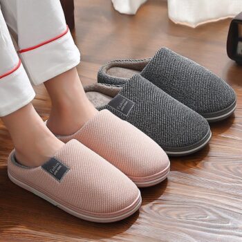 Deux paires de chaussons posées côte à côte, sur un parquet en bois, il y en a une rose et l'autre grise, l'une d'elles, la paire rose est portée, on voit une paire de jambes portant un pantalon de pyjama blanc avec un liseré rouge
