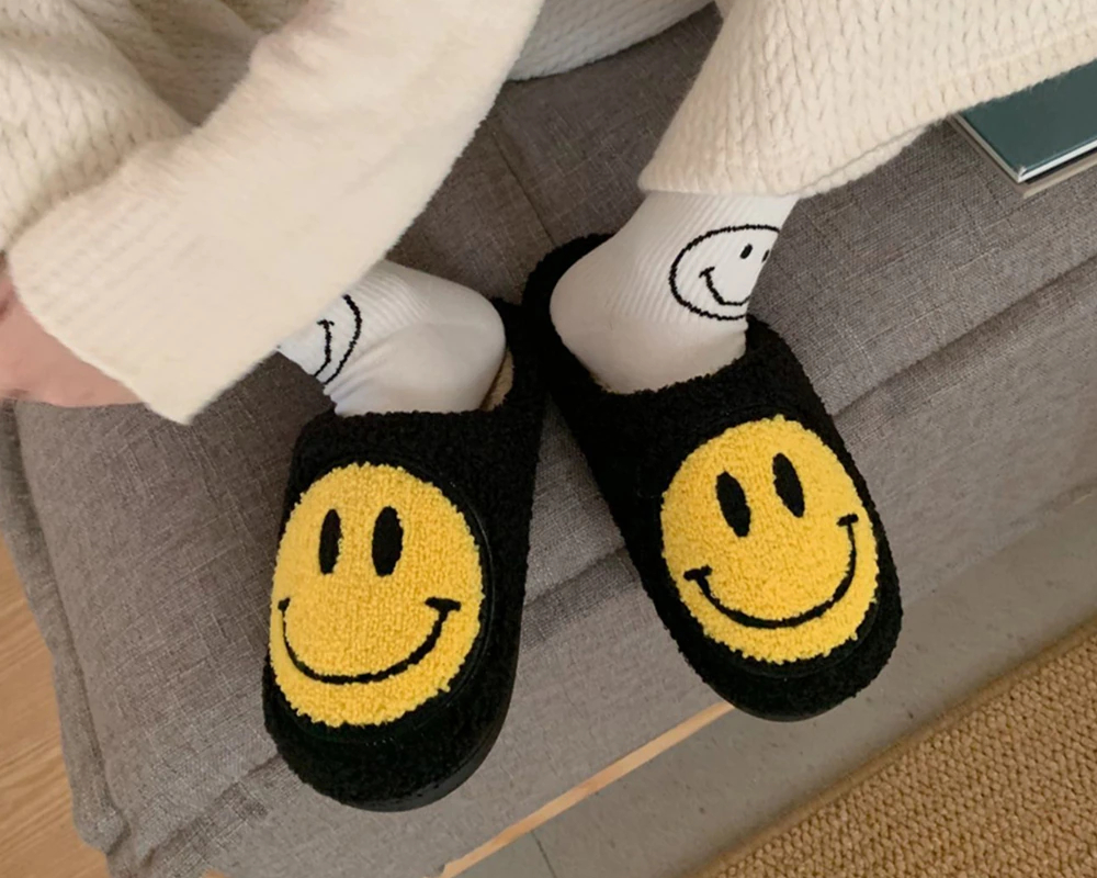 Jambe d'homme qui porte des chaussettes blanches avec un smiley noir qui sourit, et des chaussons noirs avec un smiley jaune qui sourit , ses pieds sont posé sur un tapis beige