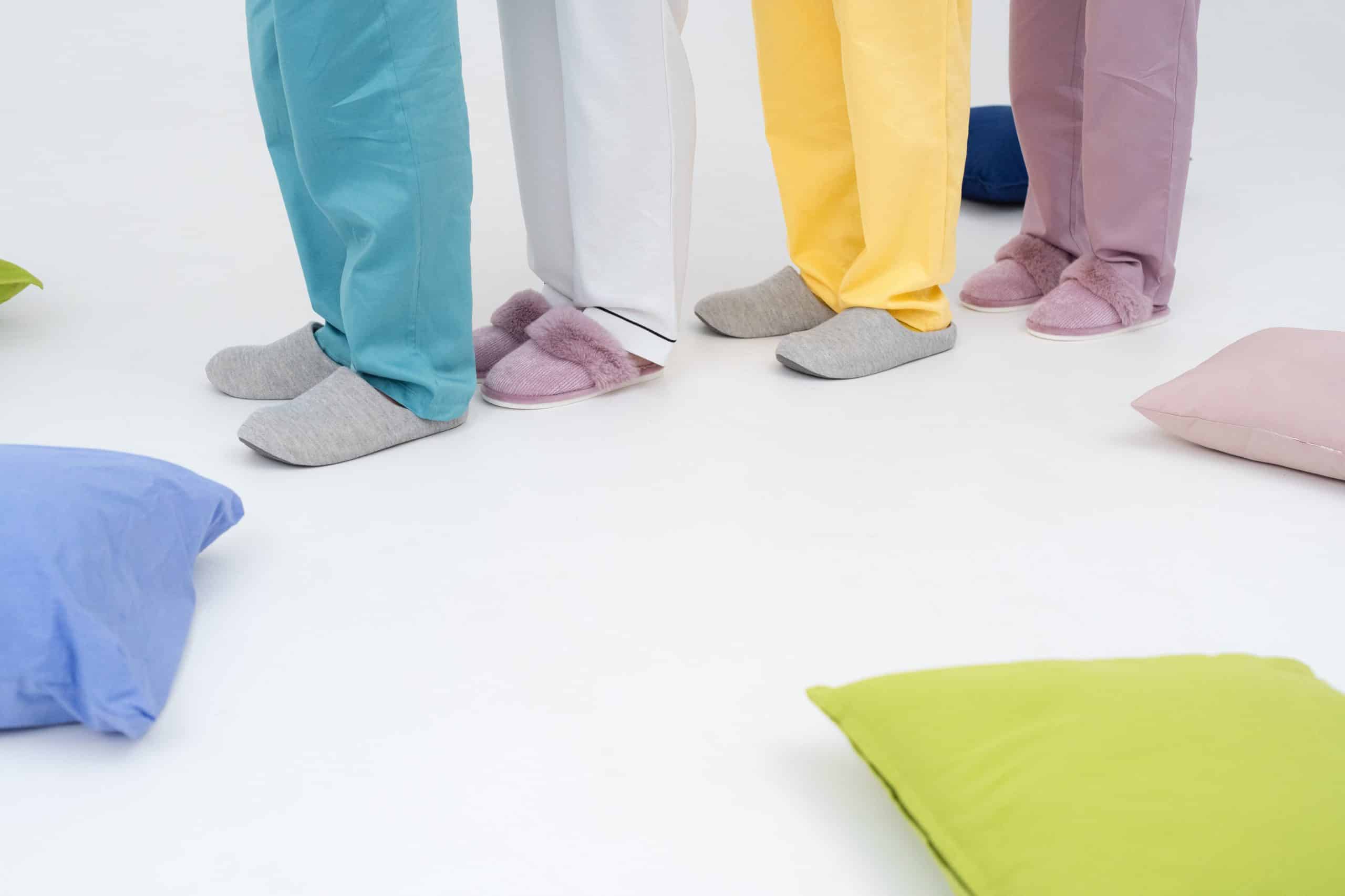 quatre personnes dont on voit les jambes à partir des genoux, se tiennt l'une derrière les autres, portent un pantalon fluide coloré (bleu, beige, jaune, bordeaux) et des chaussons, autour sont disposés 3 coussin(bleu à gauche, vert clair devant et rose à droite)