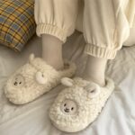 chaussons en tête de mouton portés par une personne dont on ne voit que les jambes , portant des chaussettes et un pyjama blancs, sur un lit près d'un coussin à carreau oranges
