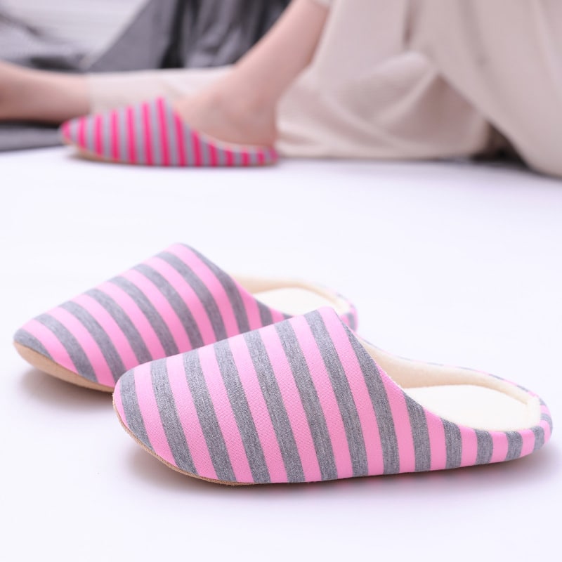 Une paire de chaussons simple rayé en gris et rose posé sur le sol blanc et quelqu'un en arrière-plan prote les mêmes chaussons