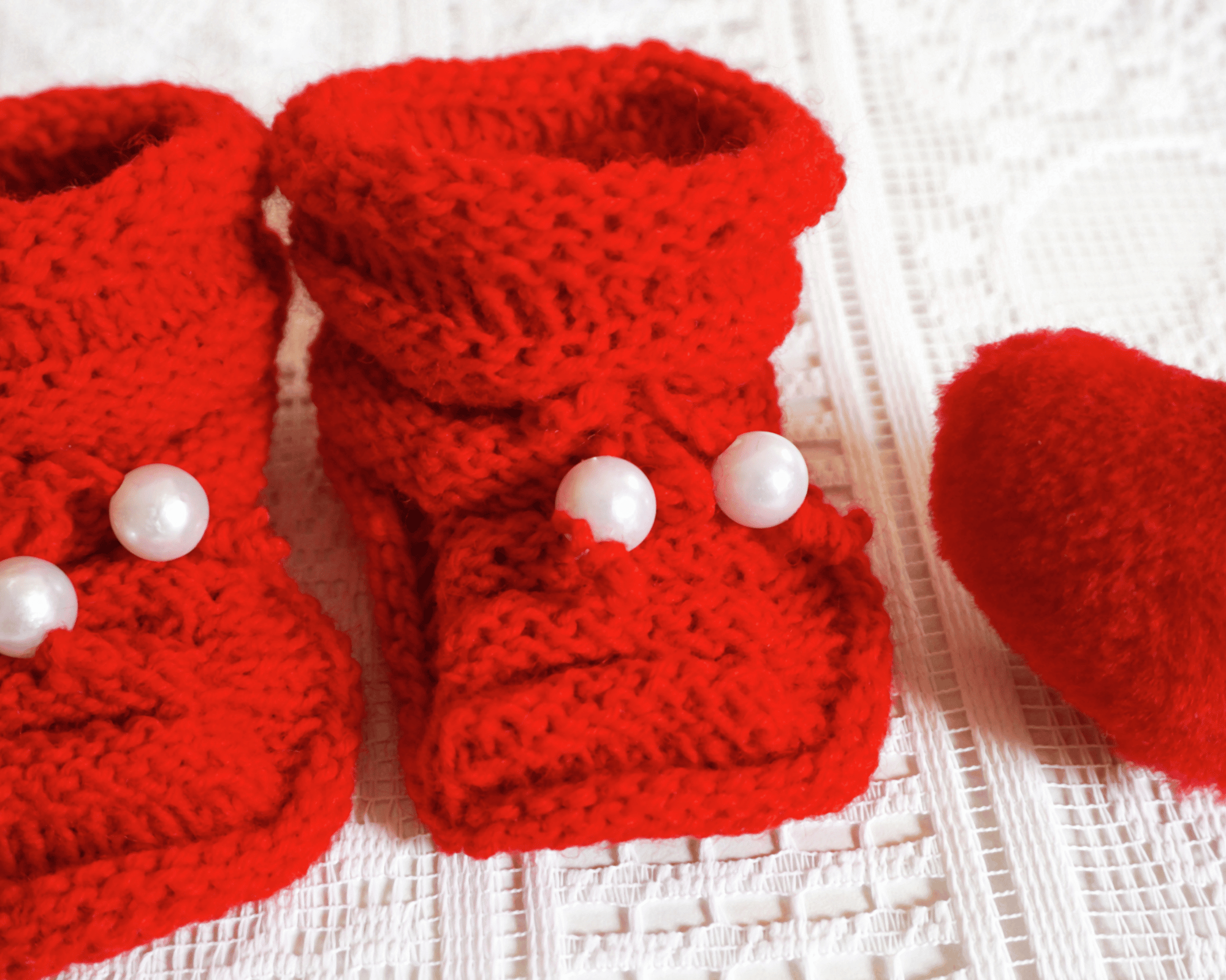 Chaussons rouges tricoté au crochet , avec 2 petites perles blanches, et avec une petite peluche en forme de cœur rouge posé à droite posé sur un tissu blanc